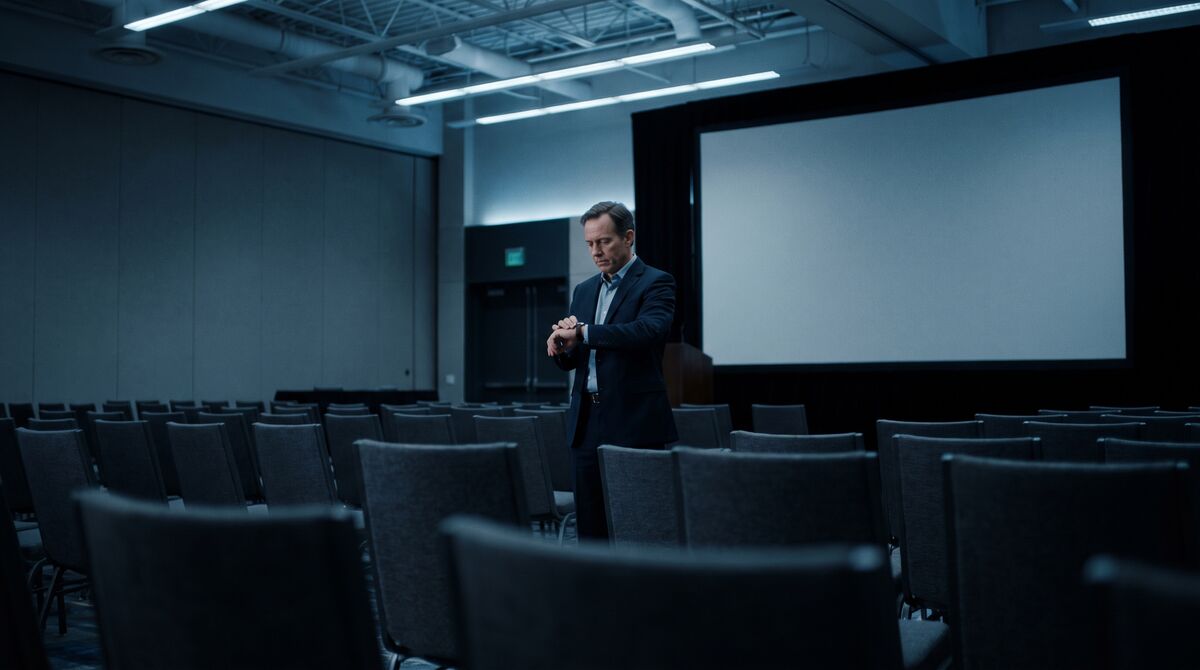 Speaker in a suit checking his watch alone in an empty conference room.